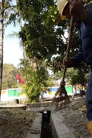 A person wearing a dark blue uniform, a light-colored hard hat, and tall yellow boots is sweeping debris from a ditch with a broom. The person is standing on the edge of the ditch and is angled away from the camera, with their arms extended as they sweep. The ditch is made of concrete and appears to have some water in it. There are trees and foliage in the background, with a red flag and a colorful wall visible further in the distance. The overall lighting suggests it is daytime.