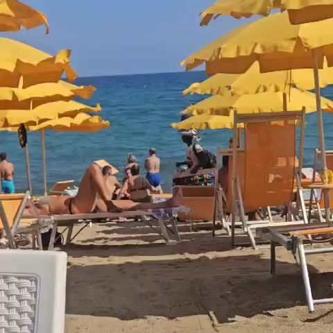 The video shows a sunny beach scene. Several yellow beach umbrellas are set up on a sandy shore. In the foreground, two lounge chairs are visible, one empty and one occupied by a person lying down. Further back, a vendor with a stack of items on their head and a backpack walks past a display of colorful items, possibly souvenirs or sunglasses. The ocean is visible in the background, with waves gently lapping the shore. Several other beachgoers are seen in the water and on the beach. The overall atmosphere is one of a relaxed summer day at the beach.