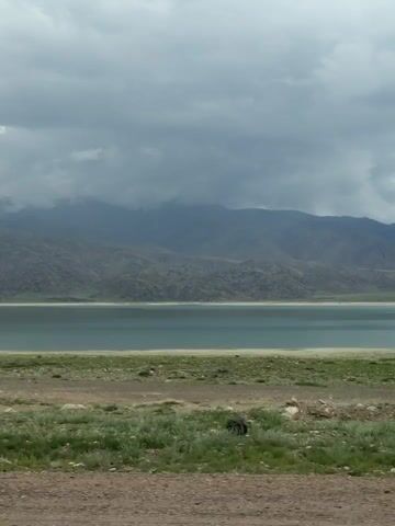The video shows a scenic view of a lake or reservoir bordered by mountains under a cloudy sky. The foreground features a dirt road and some sparse vegetation, with a concrete barrier visible along the bottom edge of the frame. The water is a calm, light blue or turquoise color, and the mountains in the background are rugged and appear to be covered in dry grass or sparse vegetation. The clouds are heavy and gray, suggesting potentially overcast or rainy weather. The camera appears to be moving, likely from a vehicle, as evidenced by the slight motion blur and the perspective shifts. There are no people or animals visible in the video. The overall impression is one of a remote, natural landscape.