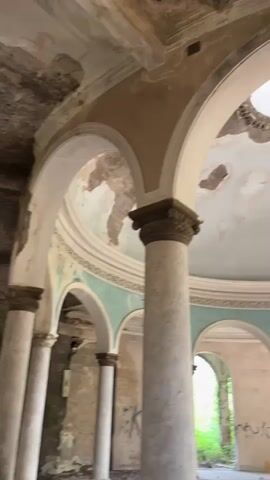 The video shows the interior of a large, circular, abandoned building. The room has a domed ceiling with a hole in the center, allowing light to filter in. The walls and ceiling are heavily damaged, with peeling paint and exposed concrete. Several large, ornate columns support the domed ceiling. Archways lead to other parts of the building, which appear to be overgrown with vegetation. Graffiti is visible on some of the columns and walls. The overall impression is one of decay and neglect.