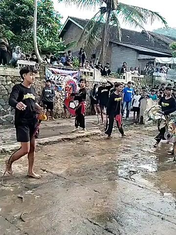 The video depicts a group of individuals participating in a cultural performance in a flooded village street. Most of the participants are dressed in black shirts, short black pants, and traditional waist wraps. Some are holding or wearing masks and other props, including what appears to be a woven fan or shield. One person is holding a whip. The street is covered in shallow water reflecting the sky. Buildings and spectators are visible in the background, and some people are filming the performance with their phones.