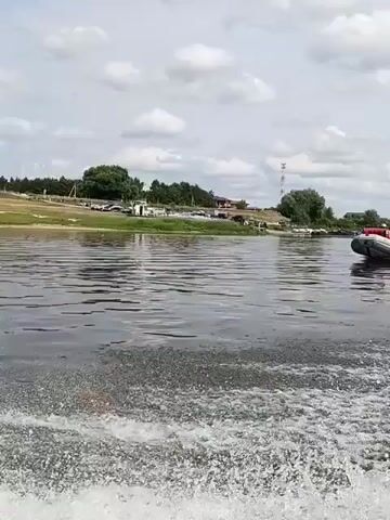 The video shows a wide body of water with a small boat on the right side of the frame moving away from the camera and leaving a wake. The boat appears to be gray and red and has at least one person in it. The water is choppy and reflects the cloudy sky. On the left side of the frame, there is a shoreline with trees and some buildings or structures. The sky is overcast with scattered clouds. The overall impression is of a recreational activity on a river or lake on a slightly hazy day.