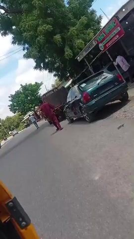 A person in a maroon outfit walks away from the camera down a road. Behind them, a truck is parked. To the right of the truck, a black car is also parked. Further down the road, other vehicles, including a three-wheeled vehicle, are visible. The scene appears to be in a tropical or subtropical climate, with lush green trees lining the road and a bright, cloudy sky. Buildings with corrugated metal roofs and open storefronts are situated along the side of the road. The road surface is asphalt.