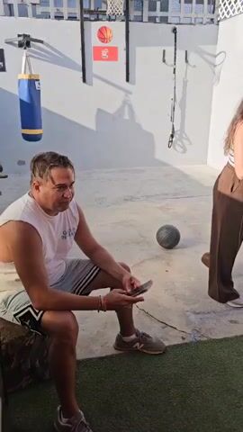 This video showcases an outdoor recreational area in Magdalena del Mar, Peru, under bright daylight.

The initial scene features a man wearing a white tank top with "COLS IN JAY" text, sitting on a tree stump and looking at his phone. A woman stands beside him, looking away from the camera. In this area, a basketball hoop with an "NBA" sign above it and a punching bag are visible, along with a black basketball on the ground.

The camera then pans to reveal a fitness and relaxation zone. This section includes green dumbbells, a black barbell with red weights, an exercise bike, and a large white beanbag chair situated on artificial turf. A gray canopy provides shade over part of the area.

Finally, the video focuses on a traditional wooden "sapito" (frog game) with metal frogs. The game board displays various scoring numbers like "1000 900 2000 900 1000," "800 700 200 700 800," "600 500 6000 500 600," and "400 300 100 400." Next to the game is an orange-cushioned wooden chair and a woven stool with a colorful blanket, set against a vibrant orange wall. The presence of shadows indicates a sunny day.