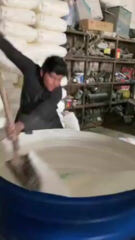A man in a black shirt stands beside a large, blue plastic drum, using a wooden paddle to stir a pale, frothy liquid inside. Behind him, there's a stack of white bags and a cluttered metal shelf filled with various items. The man works diligently, his motions indicating he is thoroughly mixing the contents of the drum. The setting appears to be a storage area or workshop.