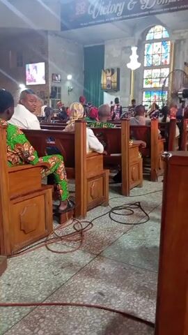 The video shows the interior of a church with people sitting in wooden pews. There are electrical cords on the floor. Sunlight streams through stained glass windows. A banner reading "Victory & Dominion" is visible at the front of the church. Some people are kneeling, while others are seated. A television screen is visible in the background.  The video pans across the rows of pews.