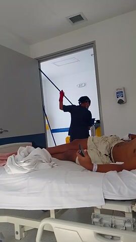 The video shows a person standing in a hospital room, wearing a blue and white striped robe, and dark gray slides. The camera angle is looking down at the person's feet. Light is shining through a window and creates a rectangular beam of light on the floor. A hospital bed and medical equipment can be seen in the background, as well as a person mopping the floor in the next room.