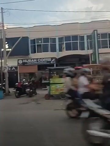 The video shows a street scene with several storefronts and vehicles. On the right side of the street, there is a building with a prominent "iPhone" sign. Next to it, another storefront displays "Hijrah Coffee". Several motorcycles are parked in front of the businesses. On the left side of the street, there is a traffic light and a few cars and motorcycles are visible. The sky is overcast, suggesting it might be late afternoon or evening. The video appears to be shot from a moving vehicle, likely a car, as indicated by the slight blur and the perspective.
