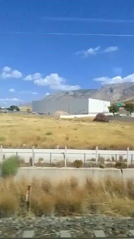 The video pans across an industrial area under a clear blue sky with scattered white clouds. In the foreground, there is dry, yellowish grass and a white fence. Beyond the fence, a winding road leads to several modern, multi-story buildings with light-colored facades and numerous windows. Some buildings appear to be warehouses or factories, with one featuring a large, light gray, curved structure. Another building has a blue glass section. Trucks are visible parked near the buildings. In the background, a mountain range with rocky, barren slopes rises against the sky. The camera movement suggests it is filming from a moving vehicle, likely a car or train, as it captures a continuous view of the landscape.