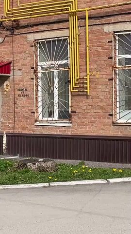 The video shows a red brick apartment building with multiple windows and doors. Yellow gas pipes run along the exterior of the building. There are benches and small flowerbeds along the building's base. A person coughs in the background. The camera pans slowly along the length of the building.