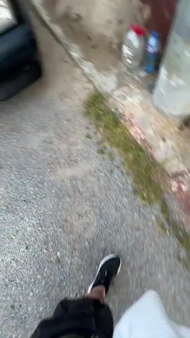 The video pans across the front of a dark-colored Mercedes-Benz car parked on a gravel surface. The car has a license plate that reads "CA 5966 PP". To the right of the car, there is a wall made of bricks and a pile of dirt. Beyond the car and the wall, there are trees and some buildings, suggesting a rural or suburban setting. The sky above is blue with some clouds. The footage is grainy and appears to be filmed with a mobile device.
