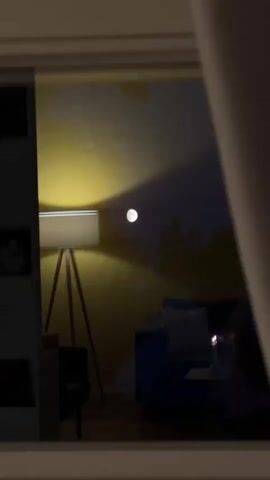 A video shows a dimly lit room through a window. A floor lamp with a white shade illuminates a yellow wall, casting a circular shadow. A bright, full moon is visible in the dark sky. A blue armchair and a table with a lit candle can be seen in the reflection. The scene appears to be a still shot from inside a room looking out a window, with the interior being dimly lit by a lamp and the exterior illuminated by moonlight.
