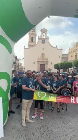 A full shot shows a group of people, approximately 50-60, gathered in an outdoor setting, presumably a street or plaza, beneath a large green and white archway. Most people are wearing matching blue t-shirts with a graphic of two hands forming a heart and the text "RUN CANCER 2023." Two adults, one male and one female, are holding a long banner that says "RUN CANCER 2023" with the same heart graphic. The banner also features text in Spanish, including "Asociacion española contra el cancer en Valencia." In the background, there is a light-colored building with a large bell tower, resembling a church or cathedral, and other buildings lining the street. The weather appears sunny with a blue sky and some clouds.