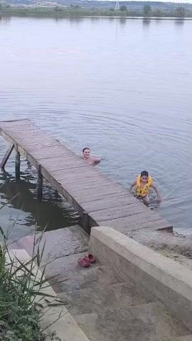 Two young males are in the water near a wooden dock. One male is closer to the camera, standing on the dock's edge, wearing a yellow life vest. The other male is in the water, swimming near the end of the dock. The water is calm, and the sky is overcast. The environment appears to be a natural body of water surrounded by vegetation and distant hills.