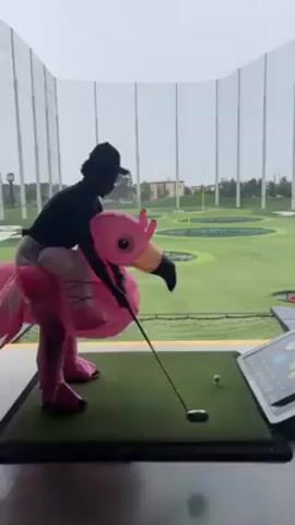 A person wearing a black cap and seated on an inflatable pink flamingo costume is holding a golf club, preparing to hit a golf ball placed on a tee on a green platform. The scene is at Topgolf, a driving range, with various circular target areas visible in the background. A digital interface displaying golfing data is visible.