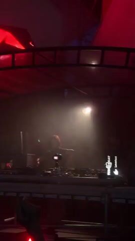 The video captures a DJ performing on a stage at night. The DJ, a person with reddish hair, is positioned behind a DJ booth filled with equipment, including turntables and a laptop. They are illuminated by spotlights, creating a dramatic effect against the dark background. The stage is decorated with red and blue lighting. In the foreground, the hands of audience members are visible, some holding cigarettes and what appear to be drinks or phones. The overall atmosphere suggests a lively music event or club setting.