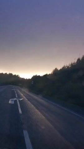 The video shows a view from inside a vehicle traveling on a wet road during what appears to be sunrise or sunset. The sky transitions from a light blue and yellow to a soft purple and pink hue. Trees line the right side of the road, and a white lane marker is visible on the asphalt. Some blurry circles, likely water droplets on the windshield, are present throughout the footage. The road curves gently to the left in the distance. There are no people or other vehicles visible.