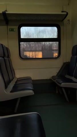 The camera shows the inside of an empty train car. There are seats on either side of a window that is showing a sunset through the trees. The interior of the train car is dated and simple. There is green flooring and white wall panels. There are posters on the wall above the window. The train is moving, though no people are visible.