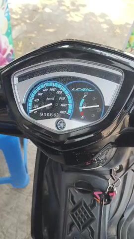 This is a first-person perspective video of a person on a motorcycle. The video begins with a close-up of the motorcycle's dashboard, showing the speedometer, fuel gauge, and a Yamaha logo. The camera then pans to the right, showing the handlebar with controls and a side mirror. The rider's hand is visible on the handlebar. The video then moves to a wider view, showing the motorcycle parked next to a road. A sign for "Mutia Juice" is prominently displayed. In the background, a person on another motorcycle is riding away. The environment appears to be a street or roadside in a sunny, possibly tropical location.