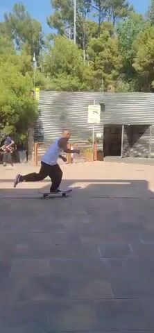 In an outdoor plaza paved with large square tiles, a young person with short hair, wearing a white tank top and dark pants, is skateboarding. They are moving from left to right in the frame, positioned in the lower center. The skateboarder appears to be in motion, possibly performing a trick as they are mid-air with their skateboard. Behind them, a building with a dark, textured facade and a light-colored, sloped roof is visible. A smaller structure with a similar roofline and signage is also present. Lush green trees and blooming purple jacaranda trees frame the scene, and there are planters with flowers and foliage in front of the buildings. In the background, a striped wall and vans are visible, suggesting an urban or park-like environment. The sky is clear and blue. Another person is seen skateboarding in the background, moving from right to left.
