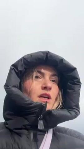 A woman in a black puffer jacket with the hood up is looking up at the sky on a cloudy day. She speaks to the camera, commenting on the weather.