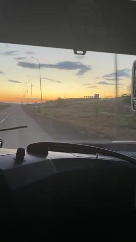 The video captures a view from inside a vehicle, likely a bus or truck, looking out through the windshield. The primary subject is the road ahead, stretching into the distance under a vibrant sunset. The sky is ablaze with hues of orange and yellow, transitioning into a soft blue as it reaches higher. Scattered clouds add texture to the atmospheric canvas.

Along the sides of the road, streetlights stand in a symmetrical pattern, some illuminated, hinting at the approach of dusk or early morning. The landscape on either side appears rural and somewhat arid, with dry vegetation and distant, low-lying hills. Construction elements, possibly bridges or overpasses, are visible in the far distance on the right.

The dashboard of the vehicle is partially visible in the foreground. A screen displays an image, and a smartphone is mounted on a stand. The overall mood conveyed is one of travel and the beauty of a natural phenomenon, the sunset, as experienced from a moving vehicle. The time of day is clearly sunset. The visible text on the vehicle's interior is "YUTONG," a brand commonly associated with buses. The location is identified as Ботақара, Kazakhstan.