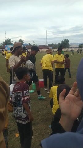The video shows a group of people, mostly young adults and children, participating in an outdoor event. They are on a grassy field, possibly a sports ground, with some buildings and palm trees visible in the background. The sky is overcast, suggesting it might be a cloudy day.

The people are dressed in casual clothing, with many wearing yellow t-shirts. Some are engaged in physical activities, while others appear to be observing or preparing. In the foreground, a child in a maroon and black jersey with the number '83' is lying on the ground. A few adults are seen standing around, and some are holding buckets. One person in a yellow t-shirt is actively interacting with a green bucket, possibly part of a game or competition.

There are several buckets, including red and green ones, scattered on the field, which could be props for a game. A person wearing a cap and a light-colored t-shirt is leaning over, seemingly engaged in an activity with a young boy. Further back, more people in yellow are gathered, and some are running or moving. A tall, slender structure, resembling a radio tower or a flag pole, is visible in the distance. There are also banners and what looks like a crowd of spectators in the background, indicating a public event or festival.

The overall atmosphere appears to be one of casual participation and perhaps friendly competition. The time of day is not explicitly stated, but the lighting suggests it's daytime. The presence of palm trees and the general environment point towards a tropical or subtropical location. Some text is visible on banners in the background, but it is too blurry to be read.