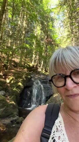 A person with blonde hair and glasses is filming a selfie video while standing in front of a waterfall in a forest. The person is wearing a white top and a black backpack. The waterfall is small and surrounded by rocks and trees. The sun is shining through the trees, creating a dappled light effect.