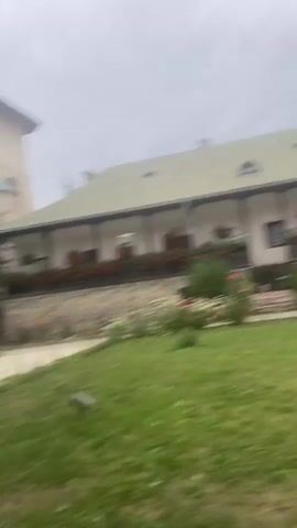 The video shows an exterior view of a building with a green roof, a long covered porch lined with red flowers, and a tall bell tower with a green cupola. The building appears to be a monastery or religious institution. There is a garden in front with various flowers and green grass. In the final clip, a woman wearing a patterned dress and a yellow jacket stands under an archway. The sky is overcast.