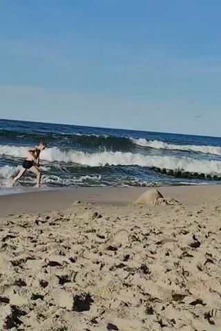 A person wearing dark shorts is standing in the shallow water at the edge of a sandy beach. They are facing the ocean, which has moderate waves. The sky is clear and blue. In the foreground, there is a pile of sand. The person appears to be interacting with the water.