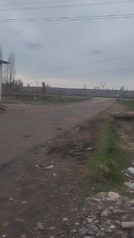 The video shows a view of a rural intersection on an overcast day. The road leading into the intersection appears unpaved and has some trash and debris along its edges. To the right of the road, there's a small ditch with some grass and litter, bordered by rocks. Power lines stretch across the sky above. In the distance, utility poles can be seen. A white truck passes on the road in the distance. A building is partially visible on the left.