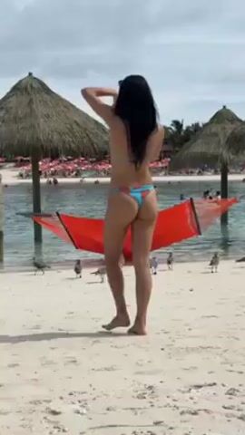 The video shows a woman with long black hair standing on a white sandy beach, wearing a bikini. She is facing away from the camera, towards a lagoon with people swimming and lounging on floats. There is a red hammock strung between two posts, and several seagulls are on the beach nearby. As the camera pans, the seagulls fly away, revealing a crowded beach with thatched umbrellas in the background.
