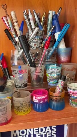 The video shows a wooden shelf cabinet displaying a collection of shot glasses, many with printed images representing different cities and countries. The shot glasses are arranged on three shelves. The bottom shelf also contains pens and markers. The overall environment suggests a hobby or collection display.