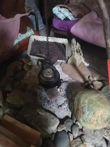 A close-up shot focuses on a makeshift cooking area. In the center, a black kettle sits on a bed of ashes and embers, suggesting a recently active fire. It is suspended by a chain from above. Surrounding the fire pit are various stones and some pieces of wood. To the left of the kettle, a white rectangular container is filled with dark, granular material, possibly coffee beans or seeds. Behind the kettle and to the right, two dried corn cobs stand upright. The background is blurry but suggests an indoor setting with blankets and pillows on what appears to be furniture, hinting at a rustic living space.