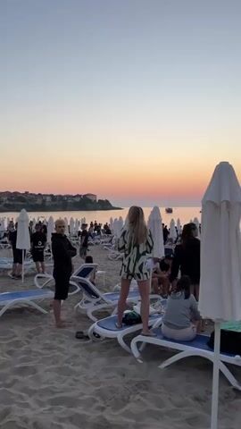 The video shows a crowded beach at sunset. Many people are sitting on lounge chairs under umbrellas. Some people are walking around, and some are sitting at tables. The ocean is in the background, and there are a few boats in the water. The sky is orange and pink, and the sun is setting. The sand is light brown. The video is shot from a low angle, looking up at the sky and the people on the beach. There are several white beach umbrellas that are closed.