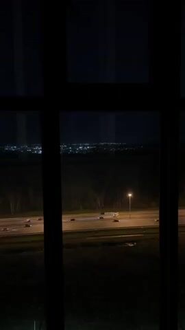 A static, close-up shot through a multi-paned window captures a night scene. Below the dark expanse of the sky, a blurred city skyline glitters with lights. A highway spans the midground, illuminated by streetlights and the headlights of passing vehicles. Between the window and the highway, a darker, indeterminate terrain can be seen.