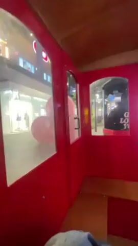 The video shows the view from inside a small, red train car moving along a sidewalk. The car has windows showing the outside. We see a shopping center with a Target store and a person walking by wearing a black hoodie with white writing and red pants. The train car has a wooden ceiling and bench seat. The camera is held by a person sitting in the train.