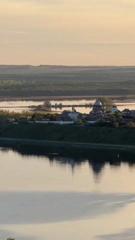 The camera pans slowly over a wide, still river, reflecting a town dominated by a cluster of white-walled churches with gold and red domes. The town is located on a green bank and surrounded by a flat, tree-covered landscape under a soft, pastel sky at sunset or sunrise. A single bird flies low over the water. The water's surface is relatively smooth, broken only by the occasional small wave. The overall impression is serene and picturesque.