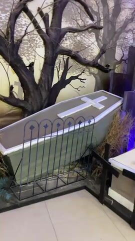 The video shows a coffin with a white cross on its lid, placed in a graveyard-themed setting. The coffin is partially obscured by a black metal fence. Behind the coffin, there is a mural depicting trees and a somber landscape. Dried grass and branches are scattered around the coffin, adding to the eerie atmosphere. The lighting appears to be dim, with hints of purple light coming from the right side of the frame. There are no people visible in the video. The scene is static, with no discernible actions or interactions occurring.