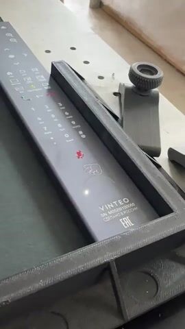 The video shows a close-up, slightly angled shot of a dark grey, rectangular object with a flat, glossy surface. The surface is labeled with white text and symbols, including the brand name "VINTEO" and a serial number "SN: MOS2BI00065." Below this, it says "СДЕЛАНО В РОССИИ" and has an "EAC" certification mark. The surface is framed by a dark, textured, 3D-printed material. Towards the right of the frame, a black, adjustable clamp-like device is visible. The background appears to be a workbench or table with a textured surface and some scattered, indistinct objects. The lighting is somewhat dim.