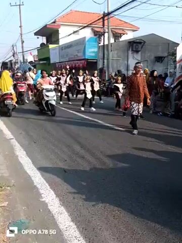 The video shows a street scene with several people and vehicles. In the foreground, a white scooter is moving left to right, with two individuals on it. To the right of the scooter, a person in traditional clothing, possibly a sarong and a striped shirt, is standing on a skateboard. Further back, a group of people in matching outfits, possibly a dance troupe or performers, are walking in procession. Some are wearing headscarves, and others are wearing distinctive headbands. There are buildings and shops lining the street, with a red and white flag visible on a pole. Other people are standing on the side of the road, observing the procession. A few motorcycles are parked along the street. The overall atmosphere appears to be festive or celebratory.