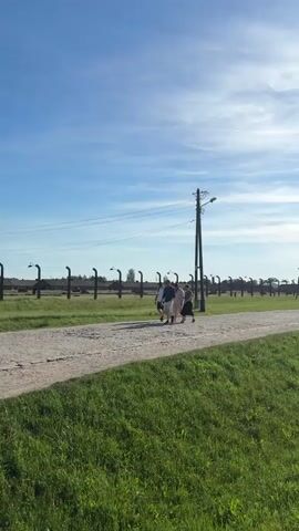 The video shows a gravel road in what appears to be a memorial site or historical location.  Three people walk along the road, heading in the same direction as other distant figures. A tall wooden guard tower stands along the road, adjacent to a barbed-wire fence.  The surrounding area is grassy. Power poles run along the road. A railway track runs parallel to the road on the right side of the frame. The sun is high in the sky, casting a bright light.  The sky is blue with scattered clouds.
