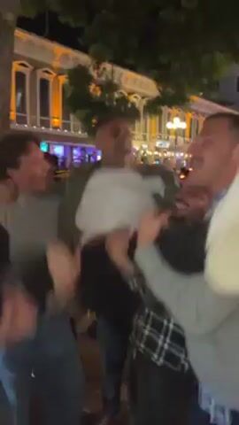 At night in what appears to be a commercial or urban area, four or five young men lift an adult man into the air, near a tree. The man being lifted wears a white shirt and black pants and appears to be laughing or shouting, as do the men lifting him. Building with orange and white lights can be seen in the background.