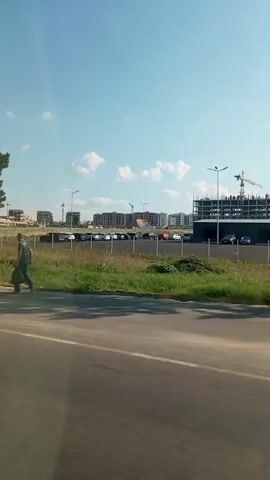 The video shows a view from a moving vehicle on a sunny day. In the foreground, there is a gray asphalt road. Beyond the road, there is a grassy area with a lone tree and a traffic sign. Further in the background, there is a parking lot with several cars, some buildings, and a construction site with a crane. The video also captures a glimpse of trees and foliage.