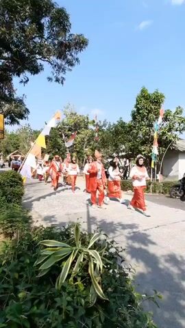 A group of people are walking in a parade down a paved path. They are wearing traditional red and white clothing, with some wearing headdresses and carrying flags. The path is lined with trees and bushes, and the sky is clear and blue. The people appear to be participating in a cultural event or celebration.