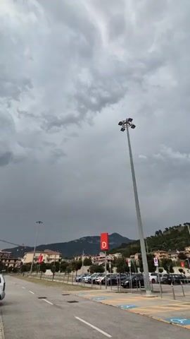 The video pans across a parking lot under a cloudy sky. In the foreground, there are several cars parked. A tall light pole with multiple lights is prominent on the right side of the frame. In the middle ground, there are more parked cars, buildings, and trees. A red sign with a white "D" is visible on a pole. In the background, mountains are visible, and the sky is filled with dark, heavy clouds, suggesting an approaching storm. The overall impression is a somewhat overcast and possibly stormy day.