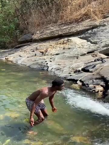 The video shows a young man standing in a shallow pool of water at the base of a small waterfall. He is wearing checkered swim trunks and is shirtless. The water is clear and the surrounding rocks are grey and tan. The man splashes some water and raises his hands, then the camera pans upward to show the waterfall cascading down the rocks. The environment appears to be a natural, outdoor setting with vegetation visible in the background.