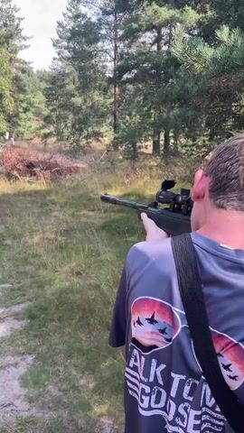 A person is aiming a rifle with a scope outdoors in a wooded area. The person is wearing a dark t-shirt with a graphic on the back that says "TALK TO ME GOOSE." The rifle is black and has a strap. The background consists of trees and grass.