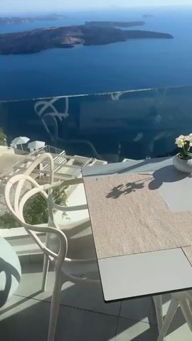 The video shows a scenic view from a restaurant or cafe with a balcony overlooking the sea. The camera pans across the scene, showcasing the expansive blue ocean with islands in the distance. Several tables and chairs, some occupied by white orchids, are set up on the balcony, suggesting an outdoor dining area. A glass railing separates the balcony from the steep drop to the sea. The video captures the sunlight glinting off the water, indicating it is likely daytime. The architecture appears to be modern and minimalist, with white structures and clean lines.