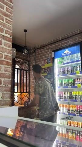 A young man wearing a camouflage t-shirt and dark shorts enters a store. He has a black fanny pack across his chest. The store appears to be a convenience store with shelves of snacks and drinks on the right side of the frame. The man walks towards the counter and then turns to face the camera. He is holding a small white object in his hands. There is an air conditioning unit mounted on the wall above the door. The walls are brick.